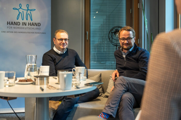 Hendrik Lünenborg und Rüdiger Ratsch-Heitmann sitzen zusammen auf einem Sofa. Im Hintergrund ist das Logo von Hand in Hand für Norddeutschland zu sehen